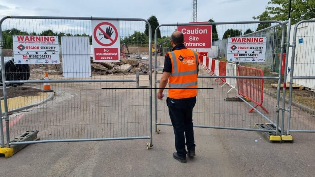 A security guard in high-visibility clothing patrolling a construction site perimeter fence.